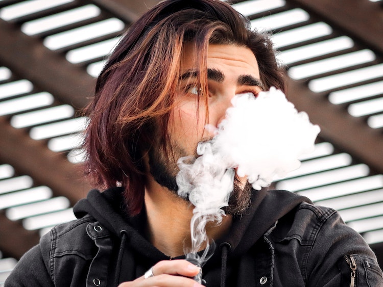 Man with long, brown hair standing outdoors exhaling cloud of vapor from vape mod