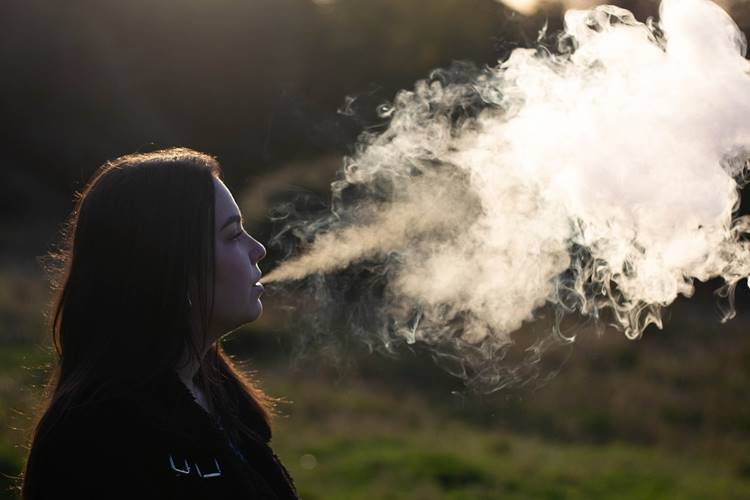 Brown-haired woman exhaling large vaporizer cloud outdoors at sunset