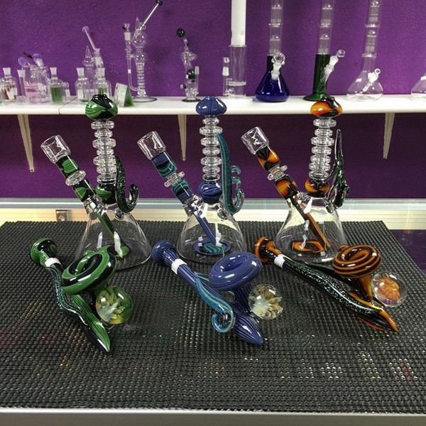 Three heady bongs in assorted colors behind row of matching heady glass pipes